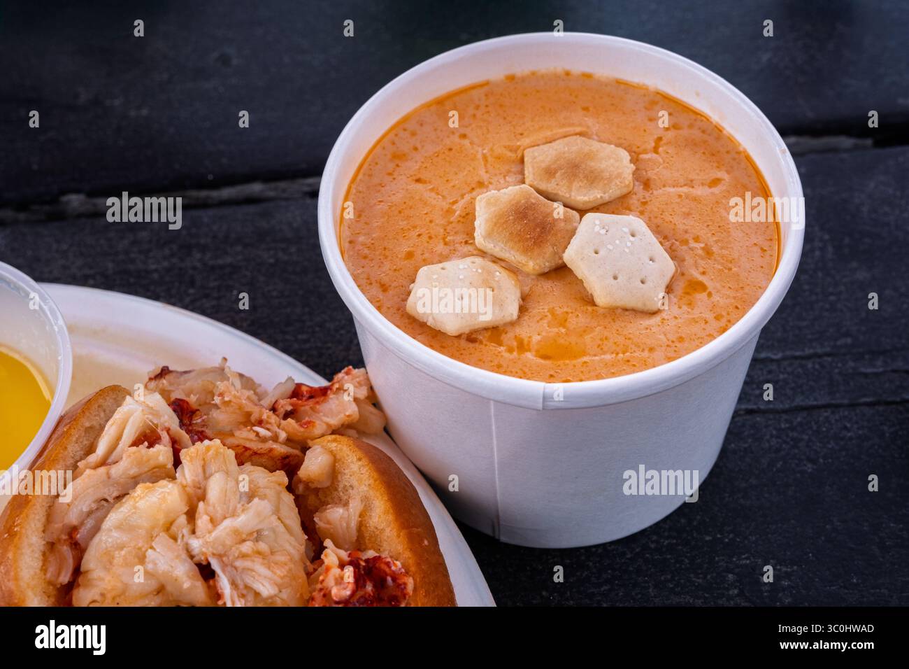 Eine Portion cremiger Hummer-Bisque mit Austerncrackern, präsentiert in einem weißen Papierbehälter auf einem rustikalen schwarzen Picknicktisch. Eine Teilansicht von Stockfoto