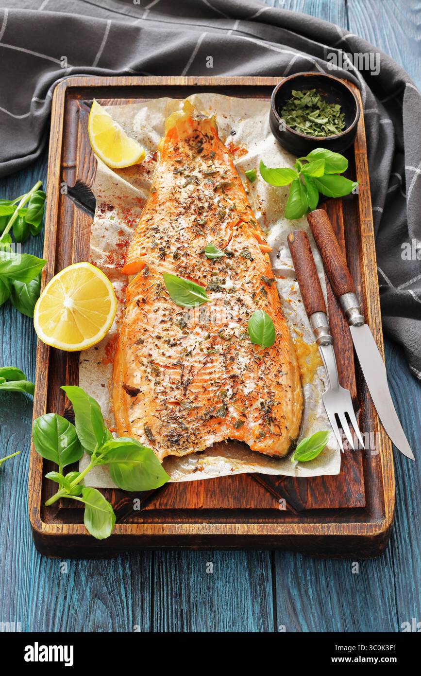 Gegrillte ganze Lachsfilets mit Haut auf einem rustikalen Holzschneidebrett mit frischem Basilikum, Zitronenscheiben, Besteck und Gewürzen auf Holztisch, vertica Stockfoto