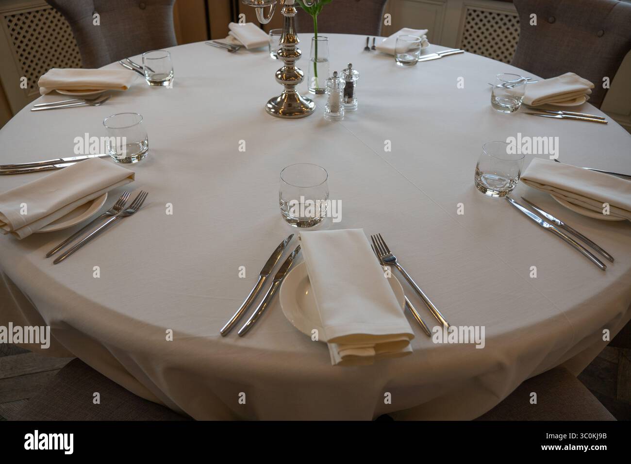 Eleganter runder Esstisch mit weißer Tischdecke, ordentlich angeordnetem Besteck, Gläsern und Servietten für sechs Gäste in eleganter Einrichtung Stockfoto