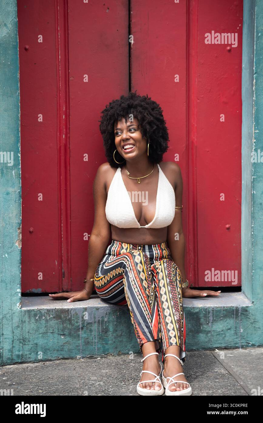 Schöne schwarze Powerfrau, die vor einer alten roten Tür sitzt. Lächelnde und befähigte Person. Brasilien Stockfoto