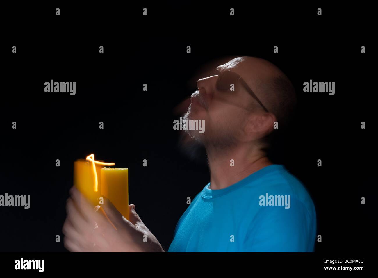 Künstlerisches Porträt eines bärtigen Glatzen-Mannes in blauem Hemd, der Bewegungsunschärfe mit brennender gelber Kerze macht. Leichte Spur. Schwarzer Hintergrund. Stockfoto