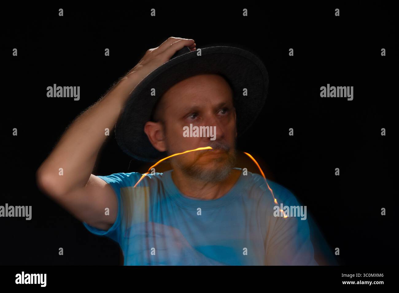 Künstlerisches Porträt eines bärtigen Glatzen-Mannes in blauem Hemd mit Hut, der mit brennender Kerze eine Lichtspur macht. Bewegungsunschärfe. Schwarzer Hintergrund. Stockfoto