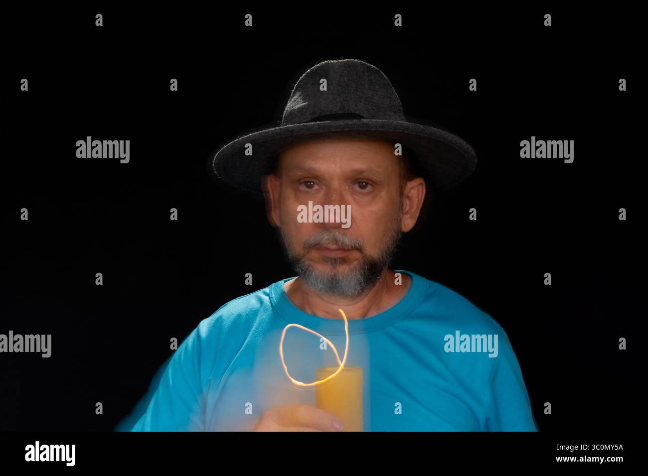 Künstlerisches Porträt eines bärtigen Glatzen-Mannes in blauem Hemd mit Hut, der mit brennender Kerze eine Lichtspur macht. Bewegungsunschärfe. Schwarzer Hintergrund. Stockfoto