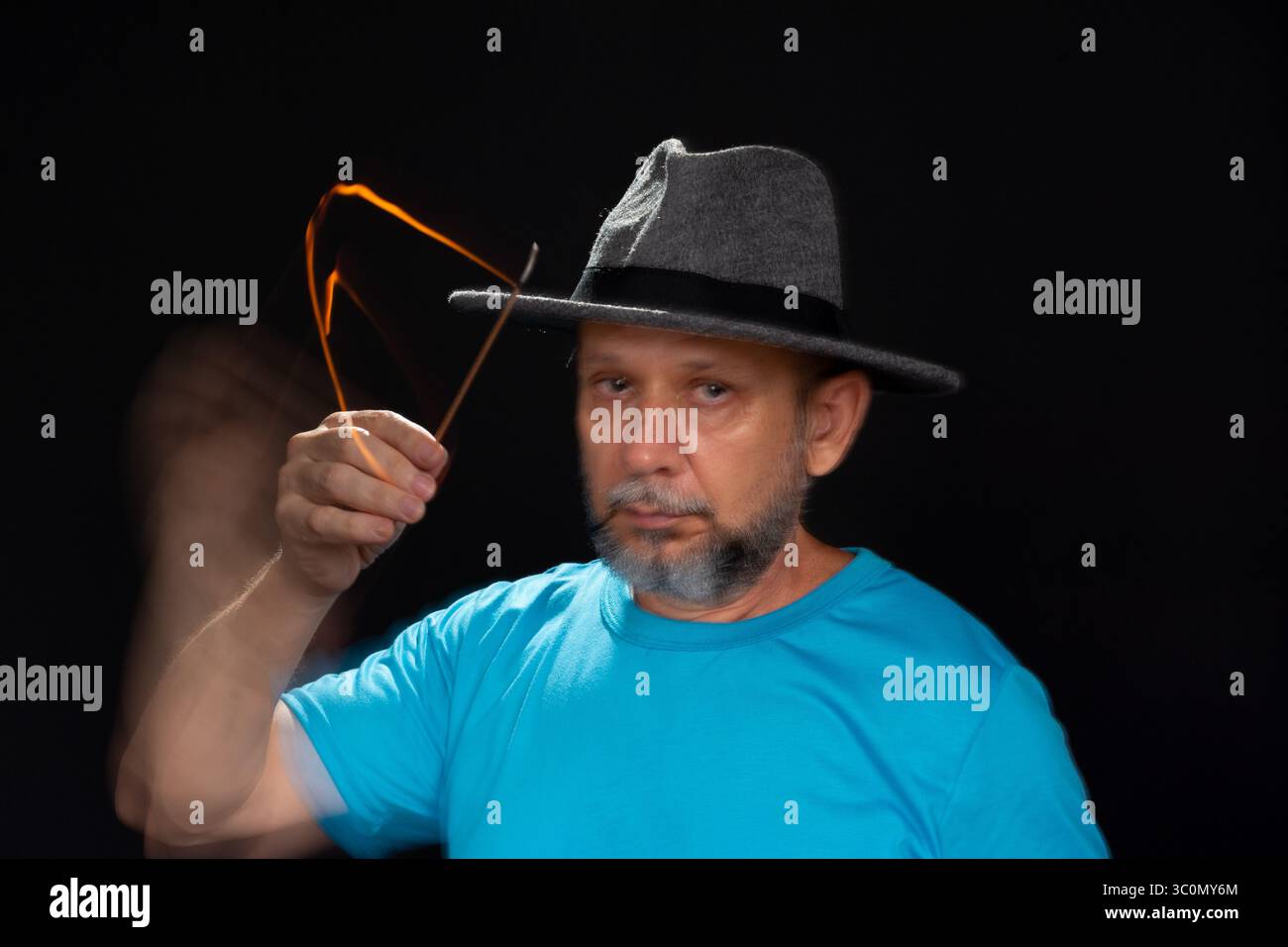 Künstlerisches Porträt eines bärtigen Glatzen-Mannes in blauem Hemd mit Hut, der mit brennendem Weihrauch eine leichte Spur macht. Bewegungsunschärfe. Schwarzer Hintergrund. Stockfoto
