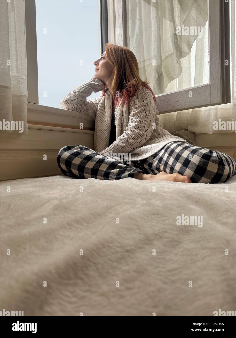 Junge Frau in gemütlicher Heimkleidung, die auf dem Bett in neutralem Schlafzimmer sitzt und nachdenklich aus dem Fenster blickt Stockfoto