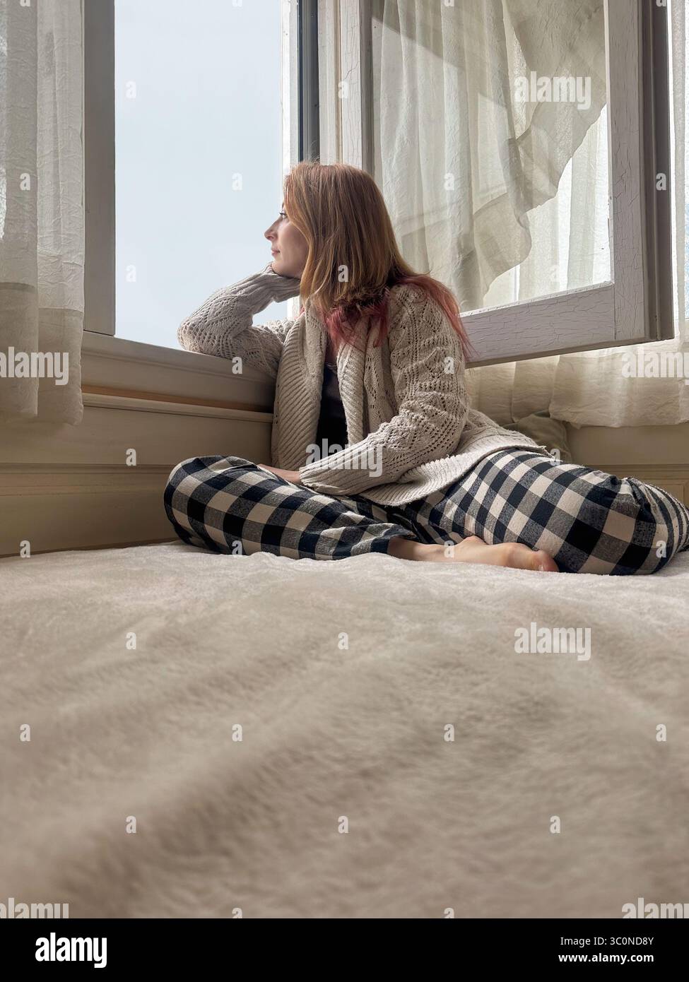 Junge Frau in gemütlicher Heimkleidung, die auf dem Bett in neutralem Schlafzimmer sitzt und nachdenklich aus dem Fenster blickt Stockfoto