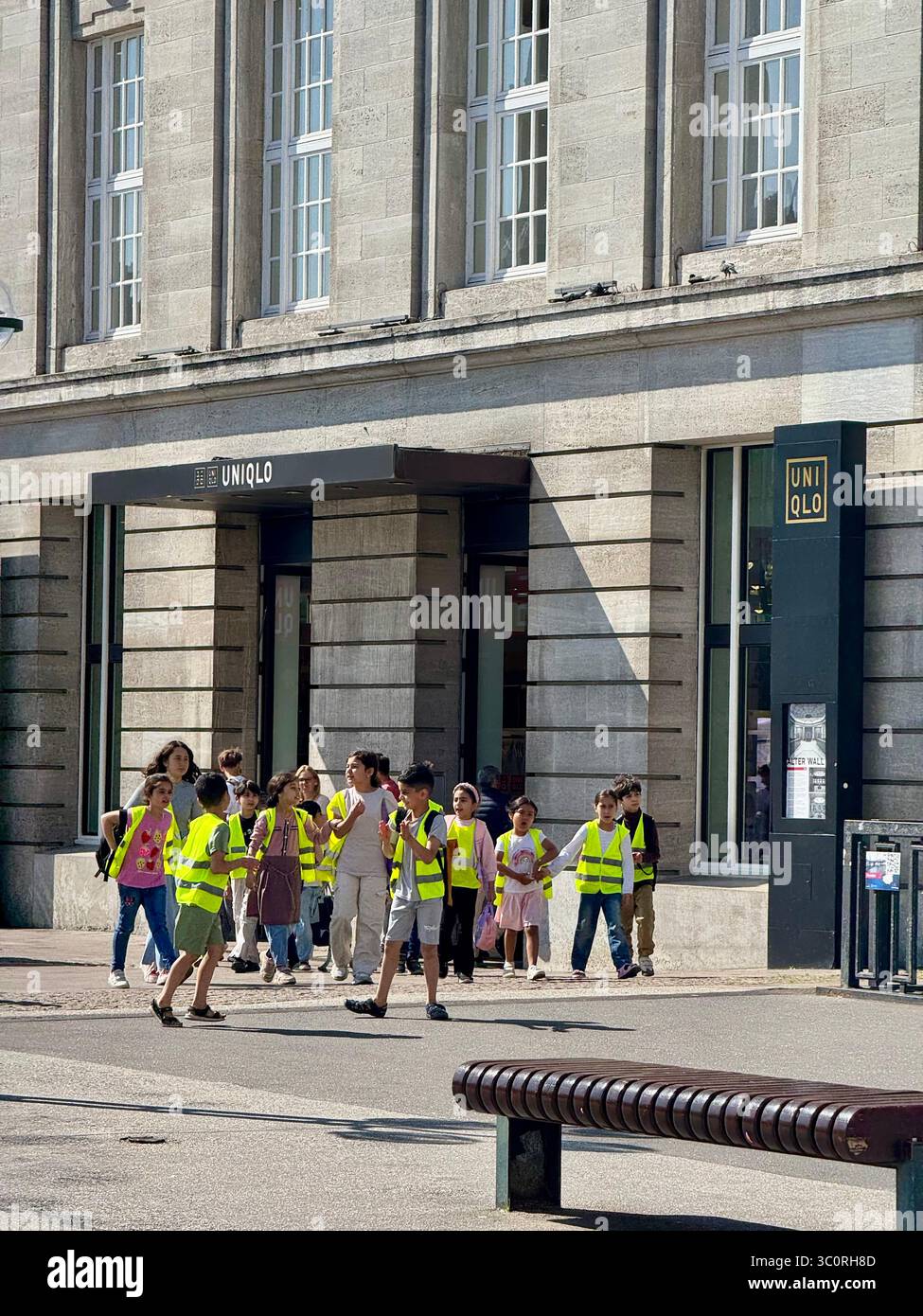 Eine Gruppe von Schulkindern und ihren erwachsenen Vorgesetzten, die alle hellgelbe Sicherheitswesten tragen, wird an einem Uniqlo-Einzelhandelsgeschäft in Hamburg vorbeilaufen sehen. - Smartphone-aufgenommenes Stockfoto