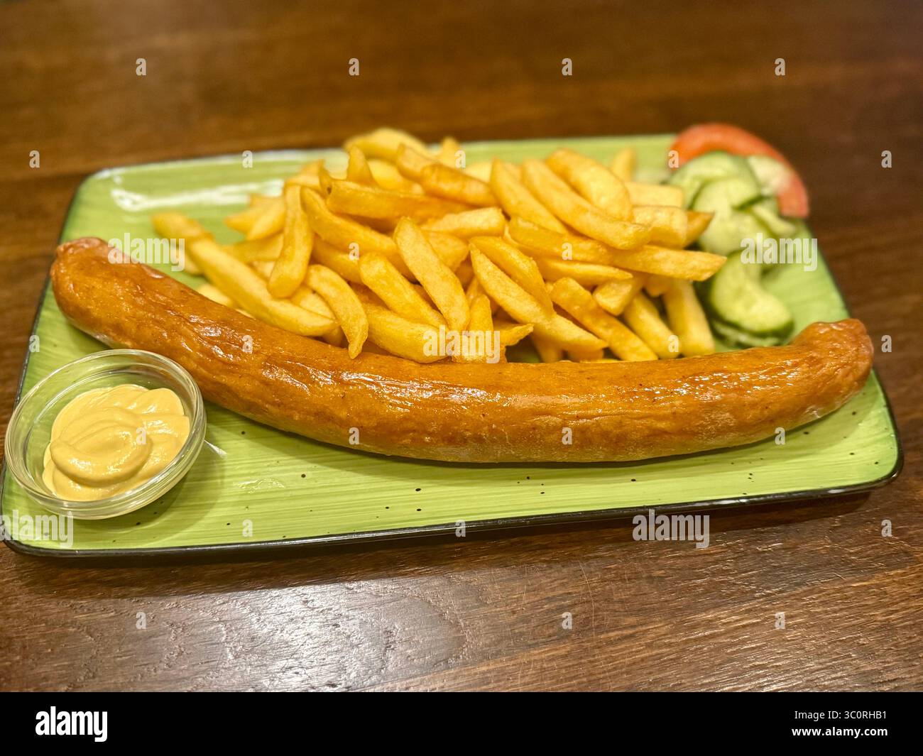 Gegrillte deutsche Wurst mit Pommes Frites, Senfdip und frischer Gurken-Tomaten-Salat, serviert auf grünem Teller in einem Hamburger Restaurant. - Smartphone-aufgenommenes Stockfoto