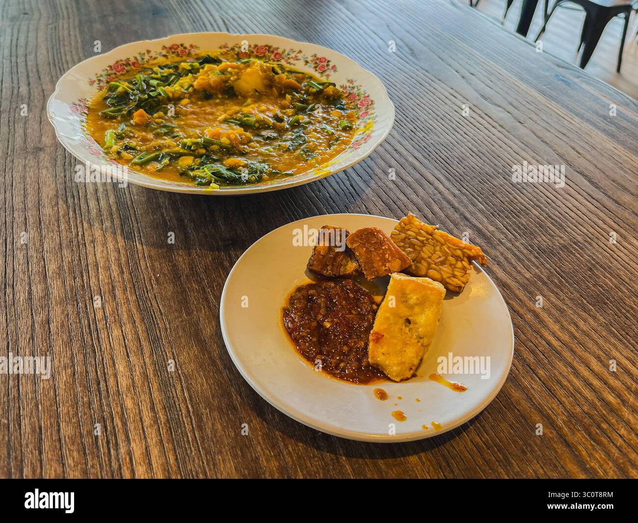 Das indonesische Gericht Bubur Manado oder Tinutuan serviert mit Sambal, gebratenem Tofu, Tempeh und knusprigen gebratenen Beilagen auf rustikalem Holztisch. Popul Stockfoto