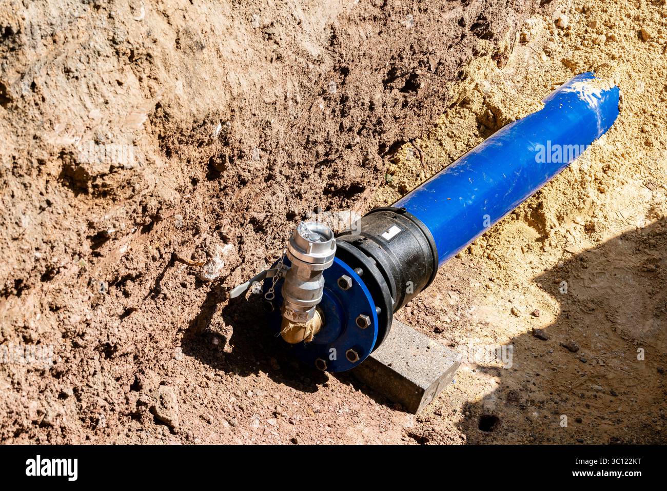 In einem Graben wird eine blaue Wasserleitung installiert. Das Gerät zeigt ein Ventil, das mit der Pipeline verbunden ist, umgeben von ausgehobenem Boden unter hellem Licht Stockfoto