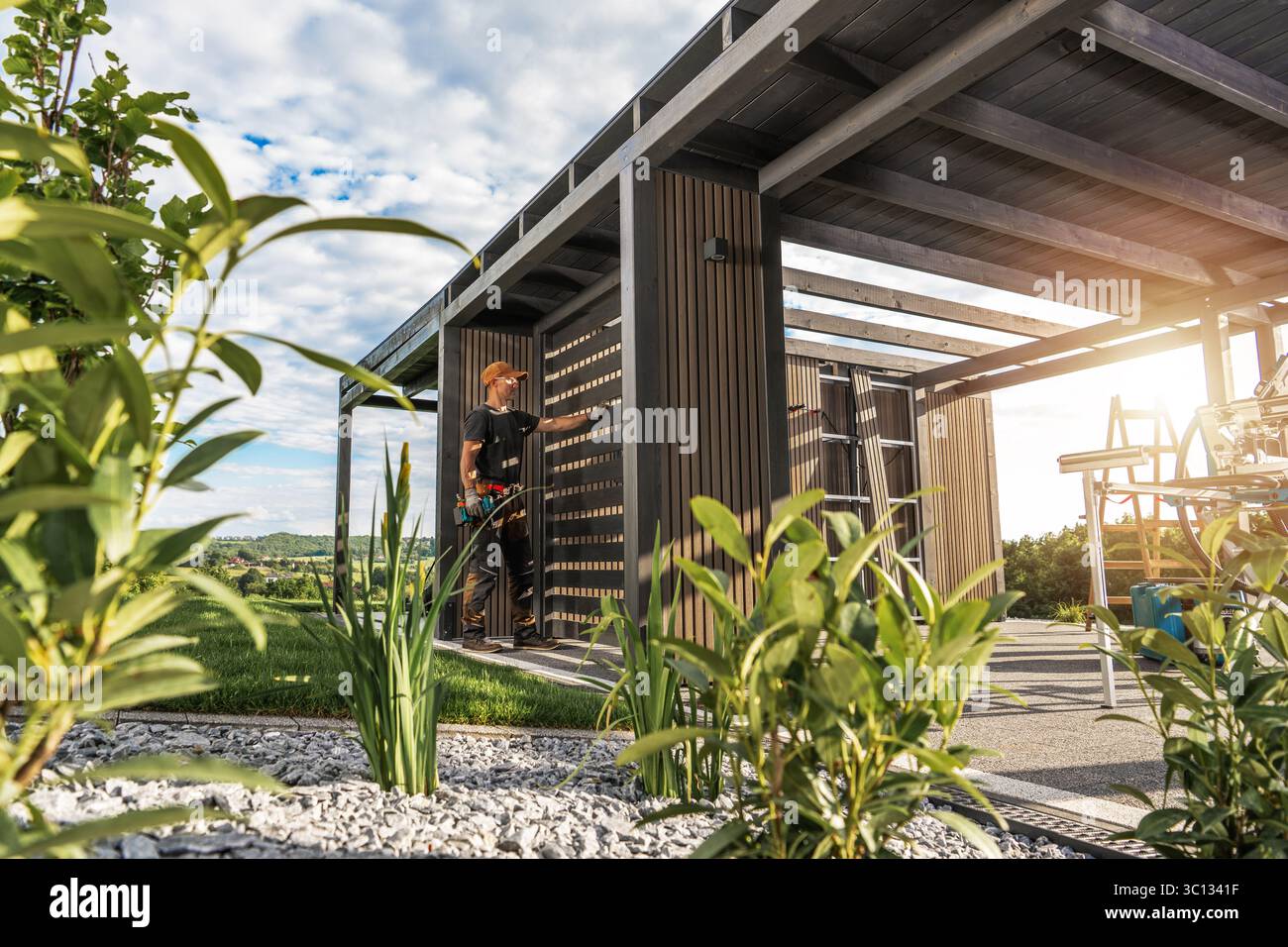Ein Arbeiter installiert am Nachmittag Holzlatten auf einer modernen Außenkonstruktion, umgeben von Grün und Werkzeugen. Die Sonne scheint im Hintern Stockfoto