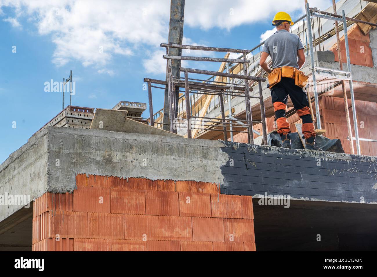 Ein Bauarbeiter steht auf einem teilweise fertiggestellten Gebäude und beobachtet die laufenden Bautätigkeiten unter klarem blauen Himmel. Stockfoto