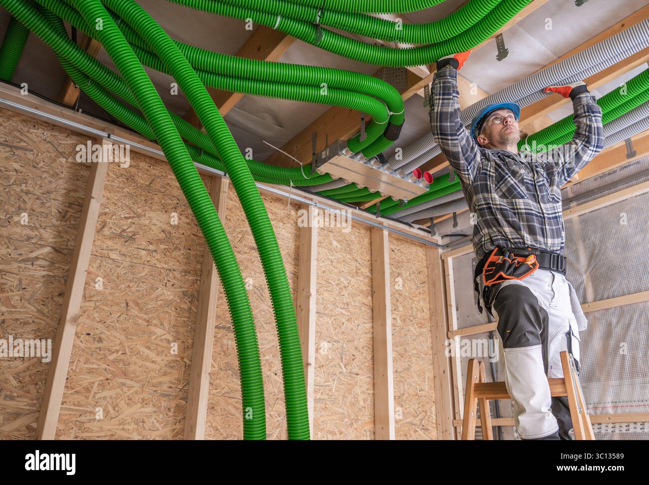 Ein Arbeiter mit Schutzhelm steht auf einer Leiter und installiert leuchtend grüne Rohrleitungen entlang der Decke einer Baustelle. Stockfoto