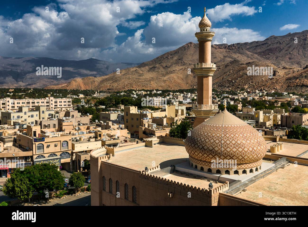 Blick auf die komplizierte Kuppel und das Minarett einer großen Moschee, die sich über der sonnenverwöhnten Stadt erhebt, eingebettet in zerklüftete Berge unter einem dynamischen Himmel, Nizwa, Oman. Stockfoto