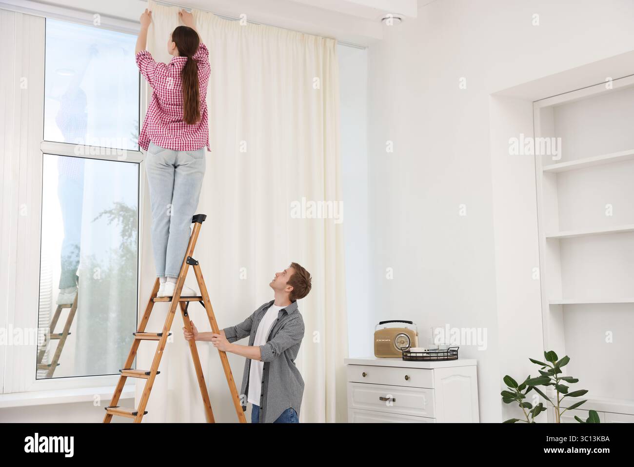 Ein Paar, das Vorhänge mit einer Leiter im Zimmer aufstellt Stockfoto