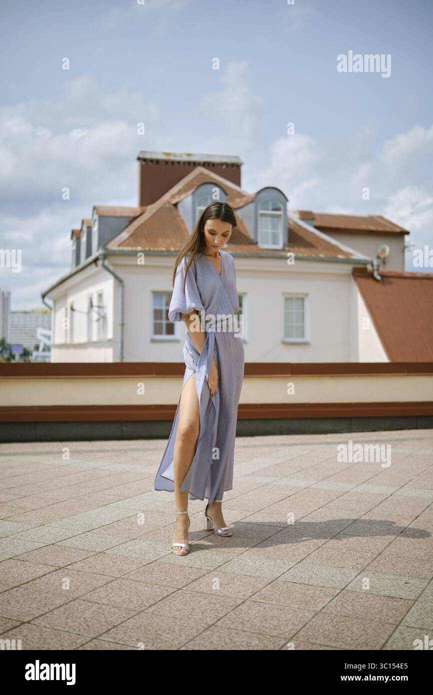 Eine stilvolle Frau steht anmutig auf dem Dach und zeigt ein fließendes blaues Kleid mit seitlichem Schlitz. Die Sonne scheint hell und unterstreicht ihr Outfit aga Stockfoto