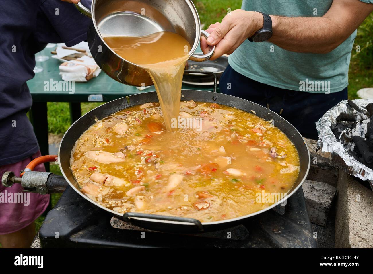 Die Köche gießen bei einem Outdoor Food Festival herzhafte Brühe in eine große Paella-Pfanne und schaffen so eine lebendige Präsentation der spanischen Küche Stockfoto