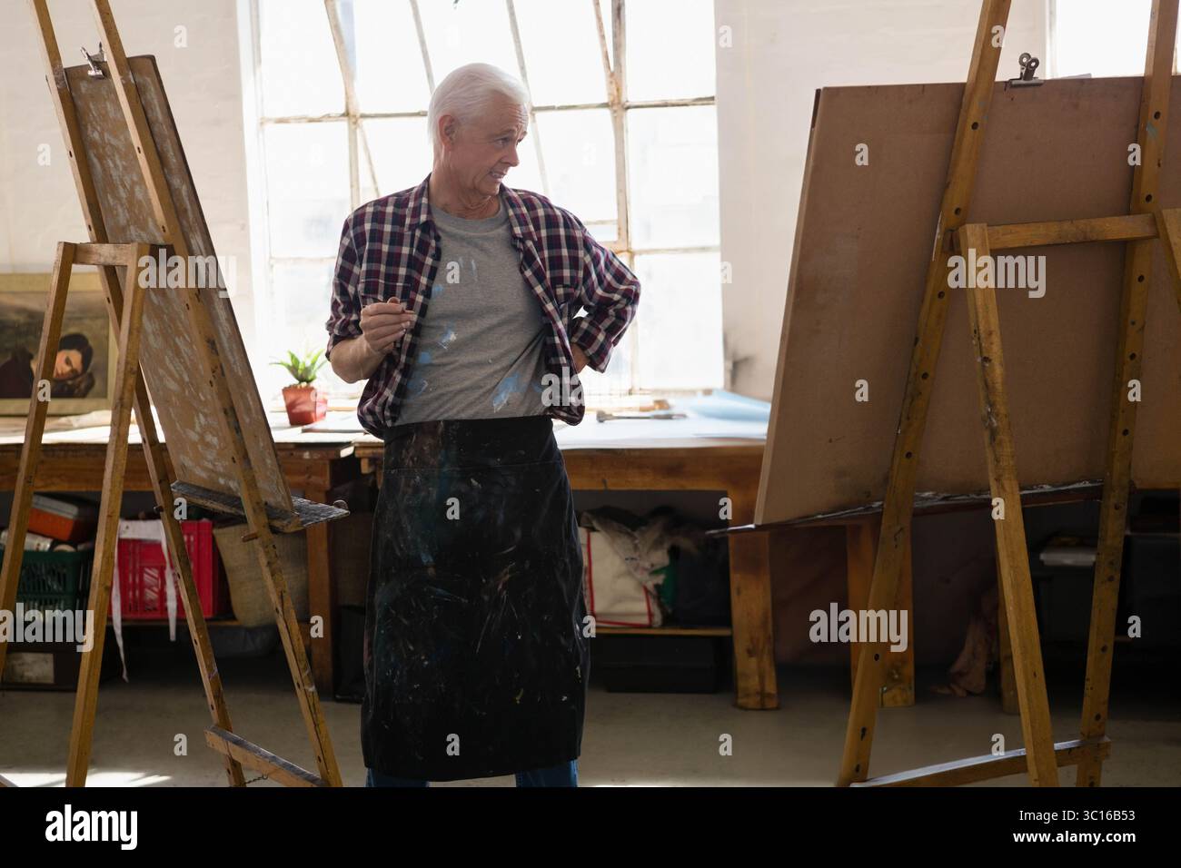 Er trug eine Schürze mit Farbflecken, ein älterer männlicher Maler hielt den Pinsel im Atelier zwischen zwei Staffeleien Stockfoto