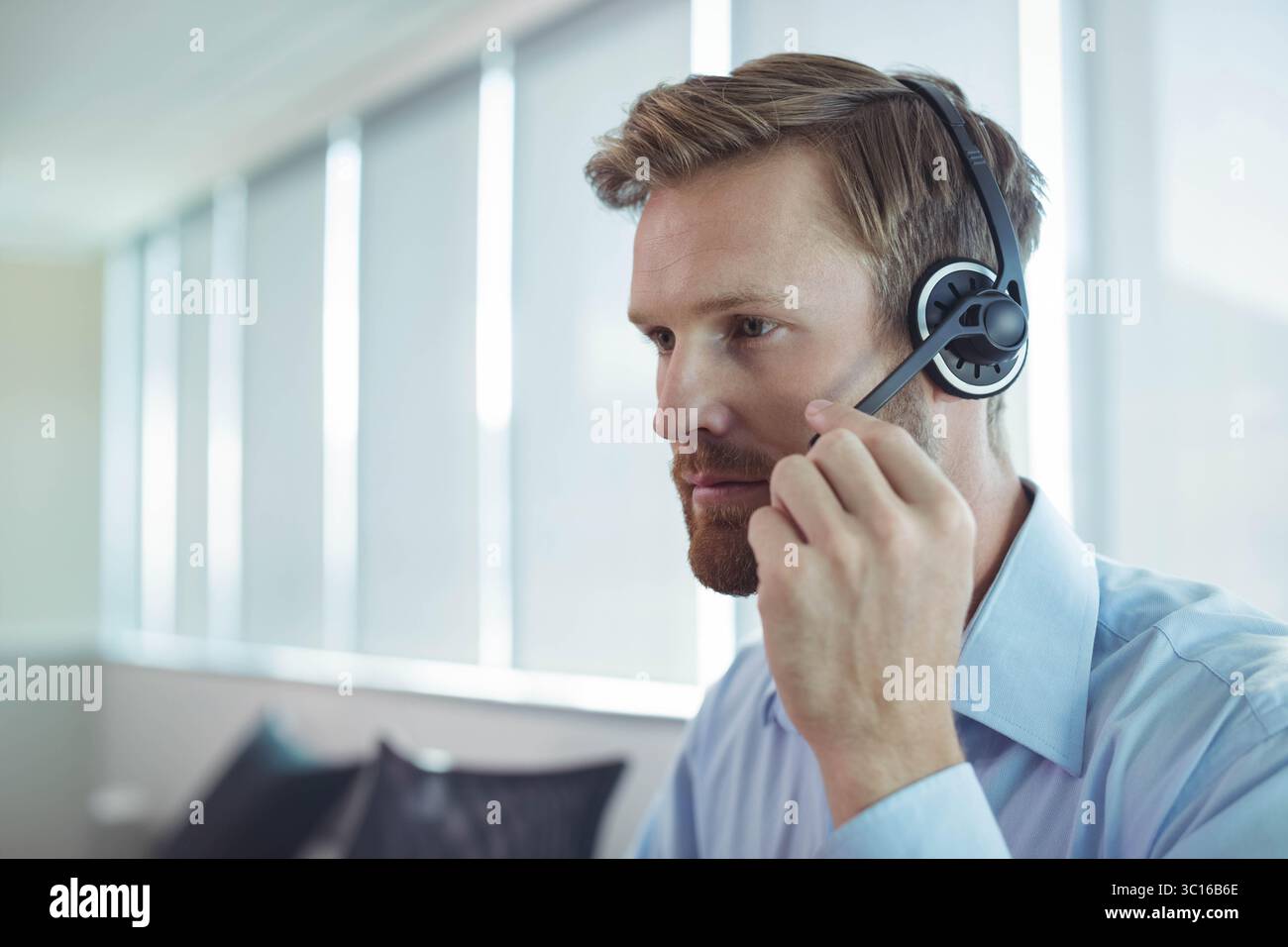Männlicher Büroangestellter, der ein Hemd trägt, das Headset-Mikrofon am Bürotisch mit Jalousien, Kopierraum anpasst Stockfoto