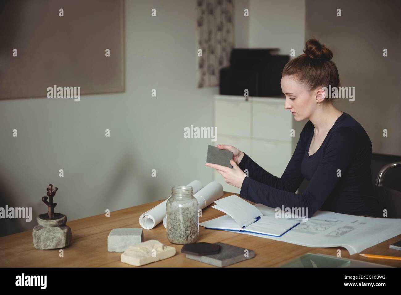 Frau inspiziert Fliesenmuster, während sie am Tisch des Designstudios sitzt, mit einem Glas mit Steinen, Kopierraum Stockfoto