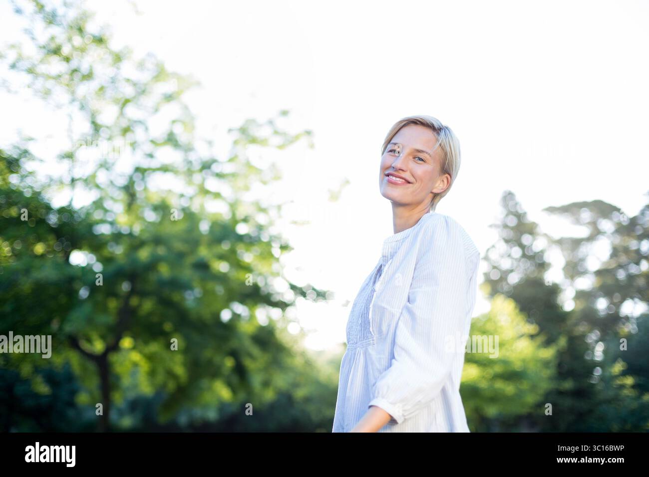 Frau in den frühen 30er Jahren, die in einem sonnigen Park zwischen Laub stand, in hellem Kleid, Kopierraum Stockfoto