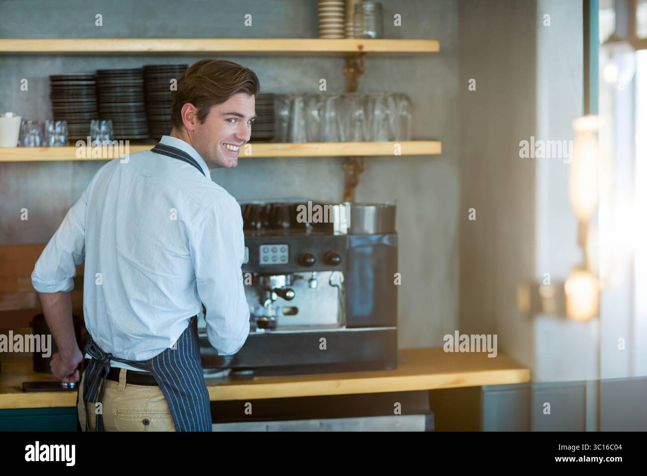 Männlicher Barista, der eine kommerzielle Espressomaschine hinter der Theke durch Regale mit Tassen betreibt Stockfoto