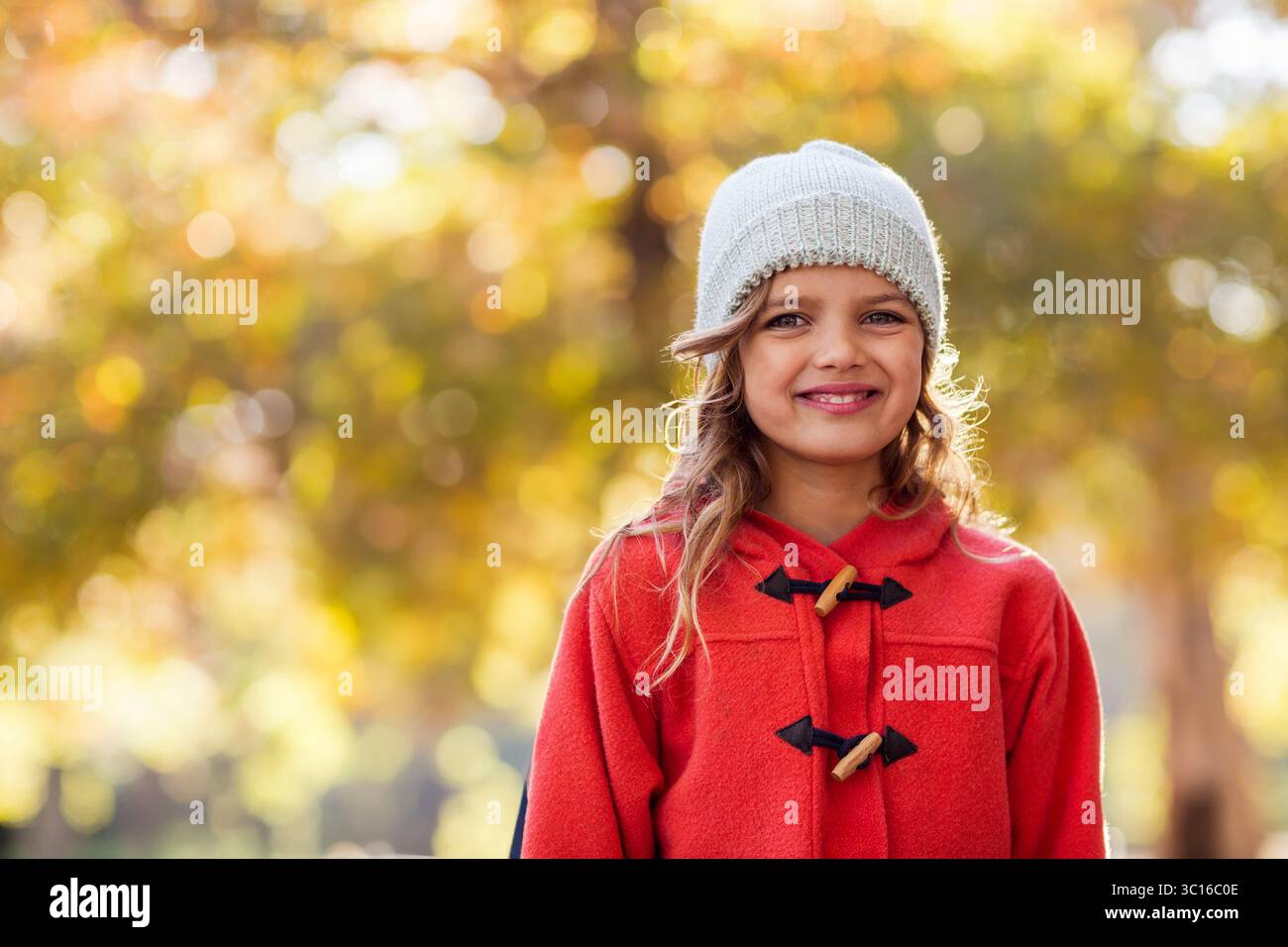 Das Kind steht im Herbstpark mit rotem Knebelmantel und hellgrauer Strickmütze Stockfoto