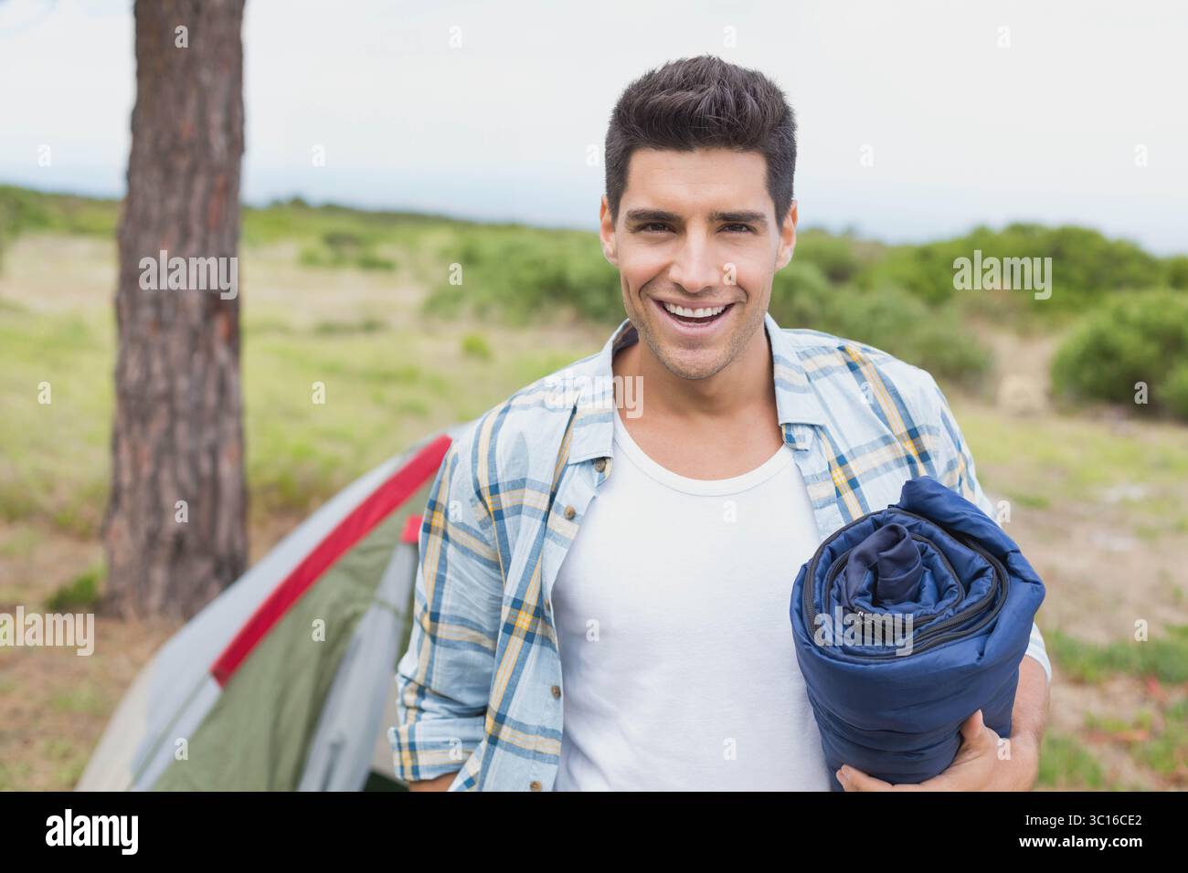 Ein Mann in den Zwanzigern, der auf dem Campingplatz steht und einen aufgerollten blauen Schlafsack neben dem grünen roten Zelt hält Stockfoto