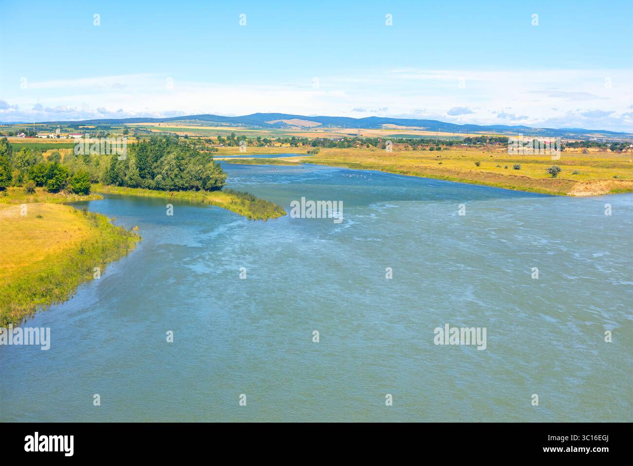 Ruhiger Wasserweg, der die Weite der umliegenden Landschaft widerspiegelt. Ein ruhiger Fluss fließt durch die grasbewachsene Ebene, die von Vegetation gesäumt ist Stockfoto