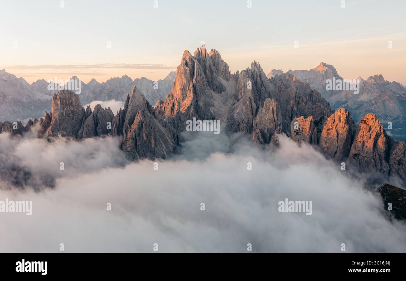 Aus der Vogelperspektive auf zerklüftete Gipfel, die durch ein Wolkenmeer durchdringen und im sanften Glanz der untergehenden Sonne getaucht sind, Parco Naturale Tre Cime, Venetien, Italien. Stockfoto