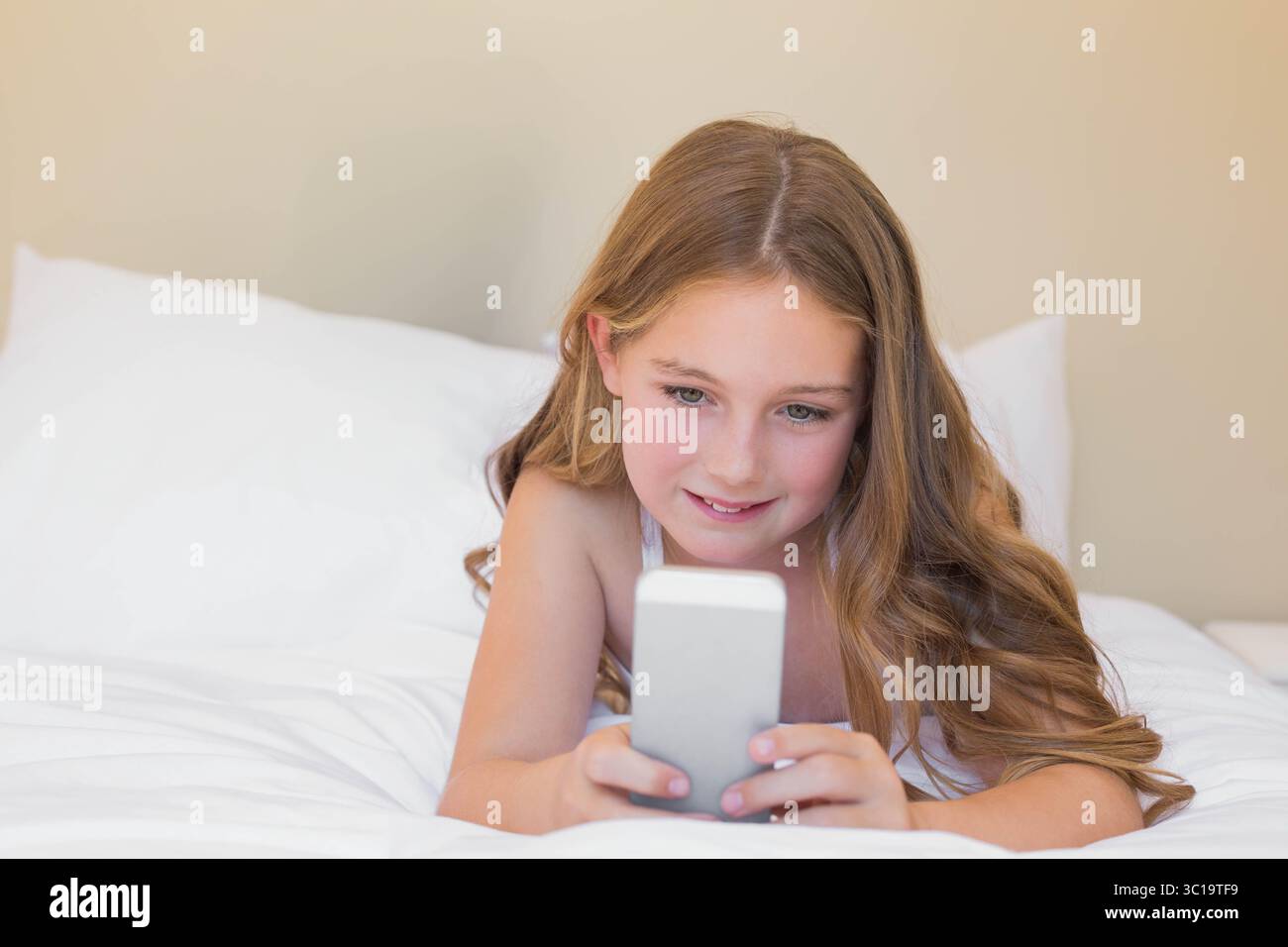 Weibliches Kind, das mit weißen Laken im Schlafzimmer auf dem Bett liegt und das Smartphone hält und auf den Bildschirm blickt Stockfoto