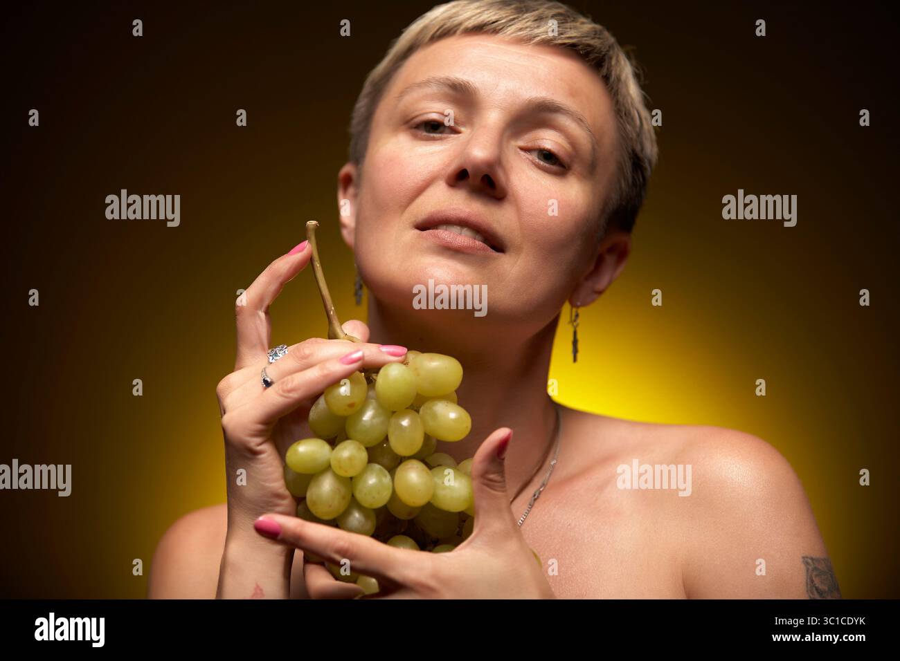 Eine Frau mit kurzen blonden Haaren hält einen Haufen grüner Trauben in den Händen. Sie blickt mit einem kontemplativen Ausdruck direkt in die Kamera. Sie Stockfoto
