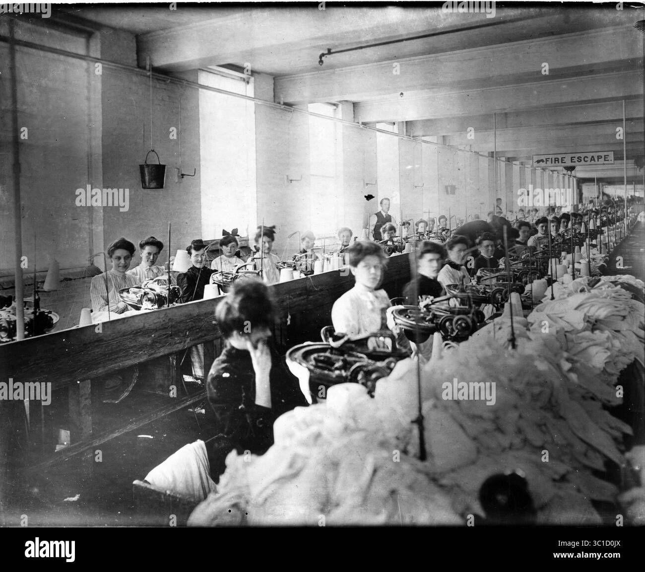 1. April 2011: Am 25. März 1911 BRACH ein Brand in der Triangle Shirtwaist Factory in New YorkÃŒÂs Greenwich Village aus. Die Arbeiter versuchten, die Treppe hinunter zu gehen, aber die Türen auf ihrem Boden waren immer verschlossen und die Feuertreppe zerknitterte bald. Im Bild: Frauen, die an Nähmaschinen in einer Fabrik sitzen, mit weißen Stoffstapeln davor. Ein Schild an der Decke auf der Rückseite steht für „Feuerflucht“ und hat eine Hand, die auf ein Fenster zeigt. (Kreditbild: Minneapolis Star Tribune/TNS via ZUMA Wire) Stockfoto