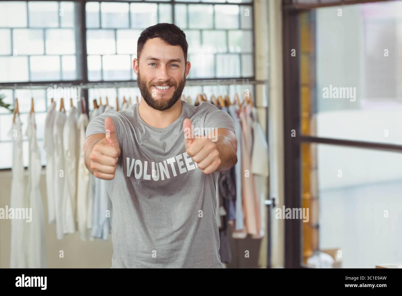 Mittelgroßer Mann, der ein freiwilliges Hemd trägt, der die Daumen nach oben zeigt, mit Hemden im Spendenbereich Stockfoto