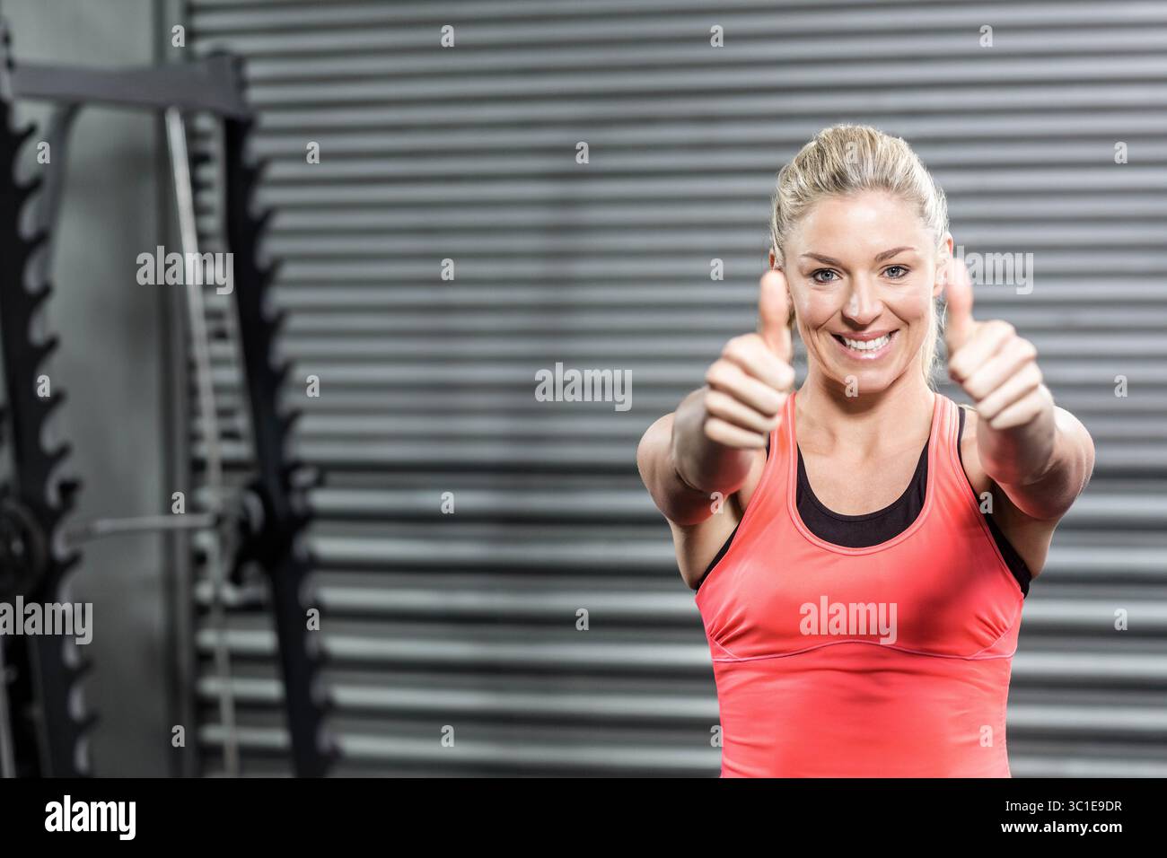 Athletische Frau lächelt und gibt die Daumen hoch im Fitnessstudio mit Langhantelablage, Hantelplatten und Auslöser Stockfoto