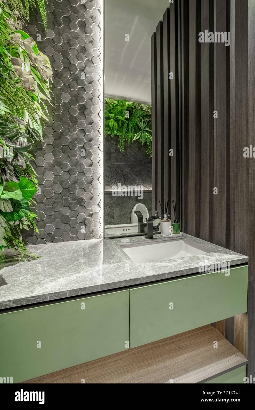 Moderner Kosmetikbereich im Bad mit grünem Schrank, grauer Marmorplatte und einem üppigen vertikalen Garten. Biophile Inneneinrichtung mit sechseckigen Fliesen und Spiegel. Stockfoto