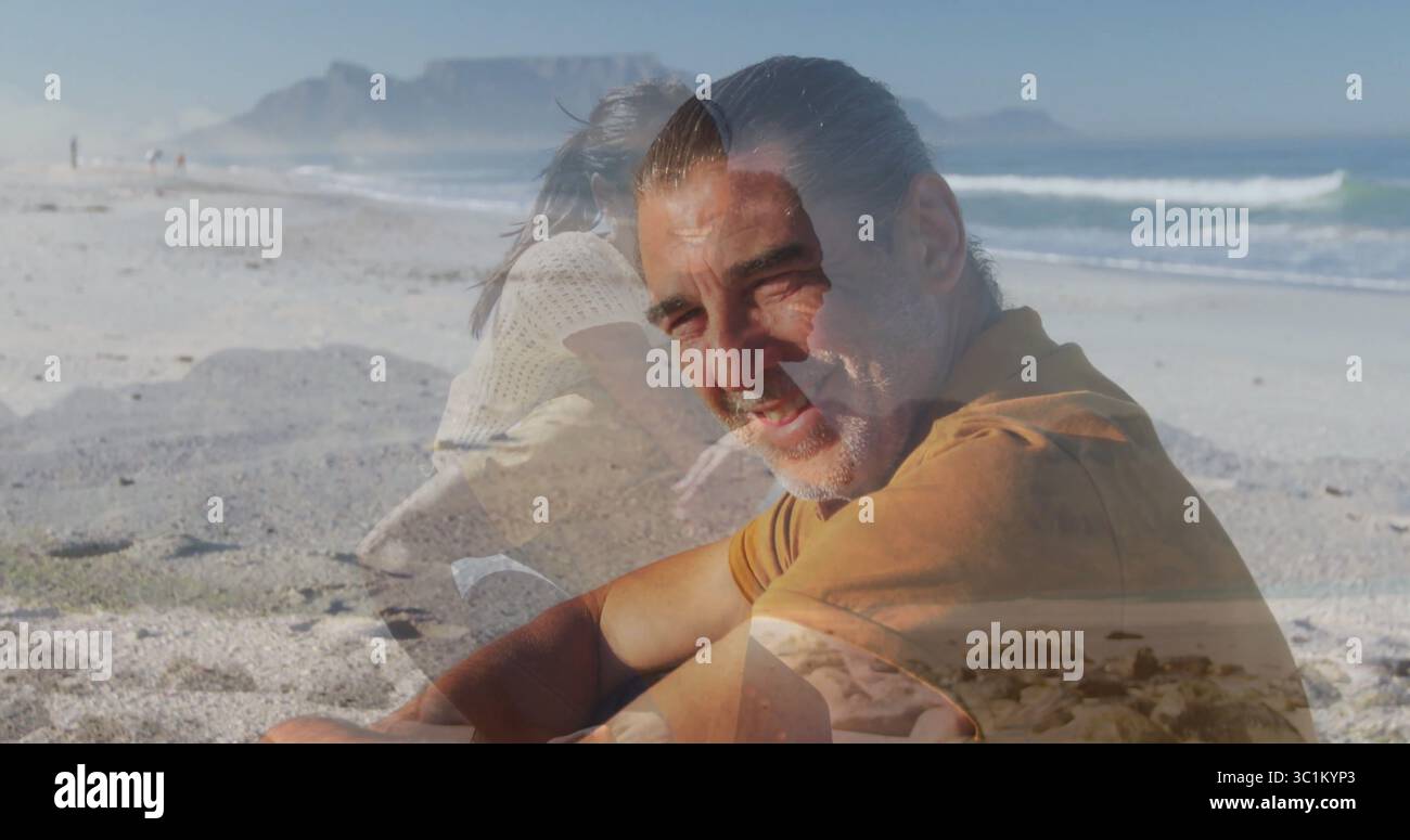 Ein lächelnder reifer Mann mit Rosthemd und Hose, der am Sandstrand sitzt, mit Meereswellen Stockfoto