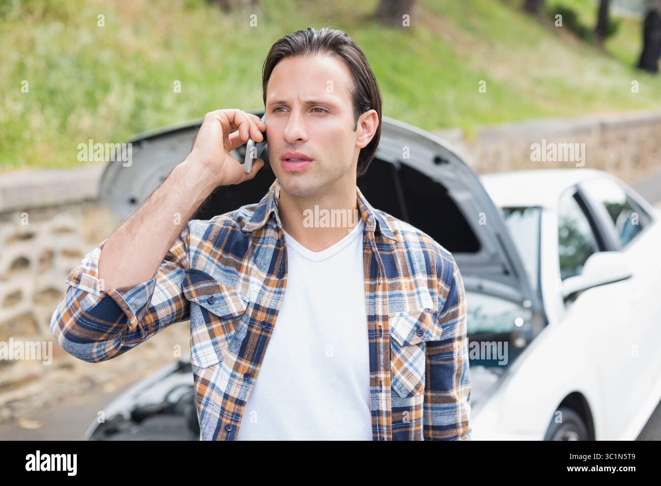 Mann, der ein Smartphone hält und am Straßenrand Hilfe ruft, neben einer weißen Limousine mit offener Haube an einer niedrigen Steinmauer Stockfoto