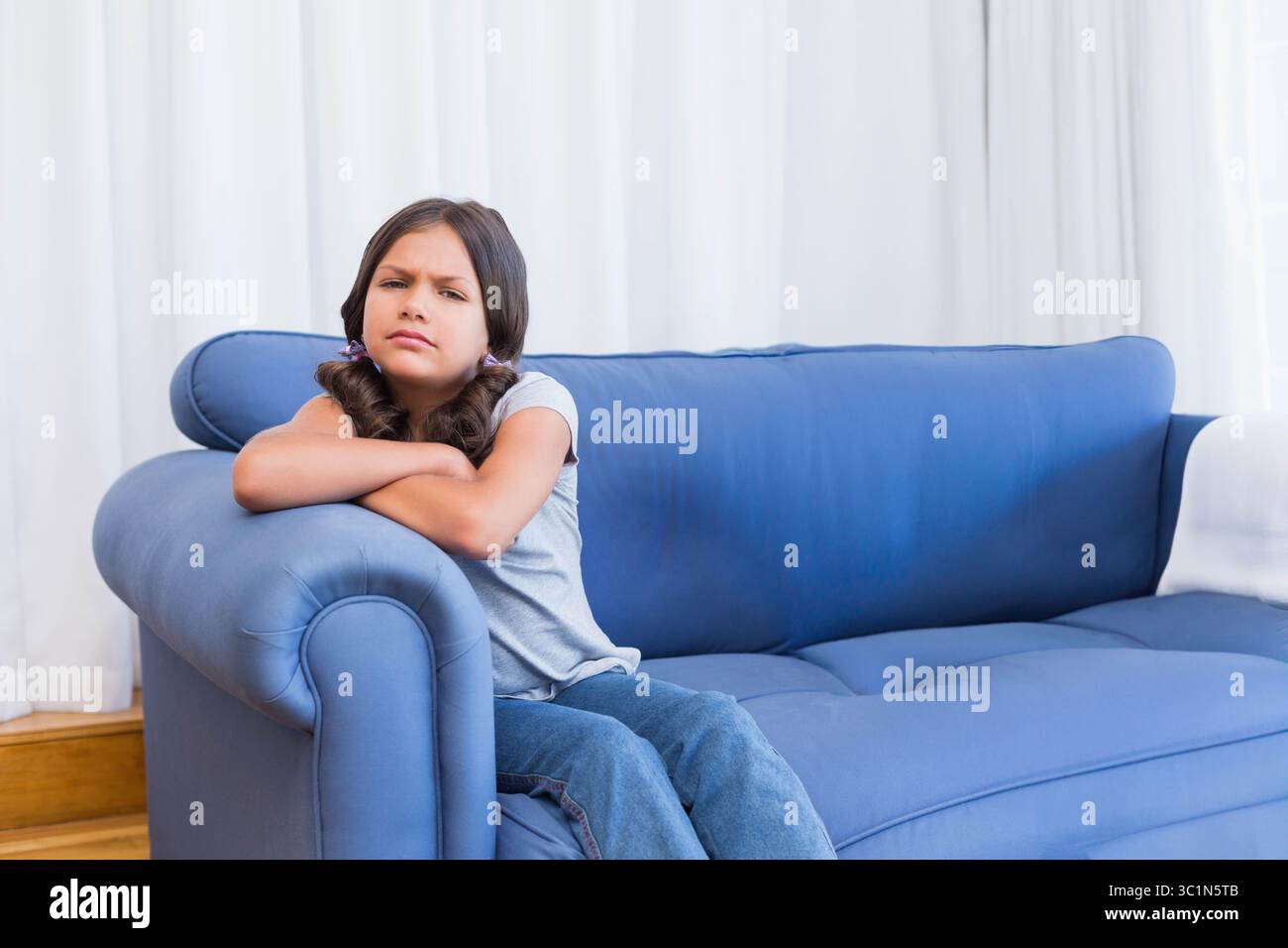 Mädchen im Schulalter, das sich im Wohnzimmer mit weißen Vorhängen auf einem blauen Sofaarm lehnt, Kopierraum Stockfoto