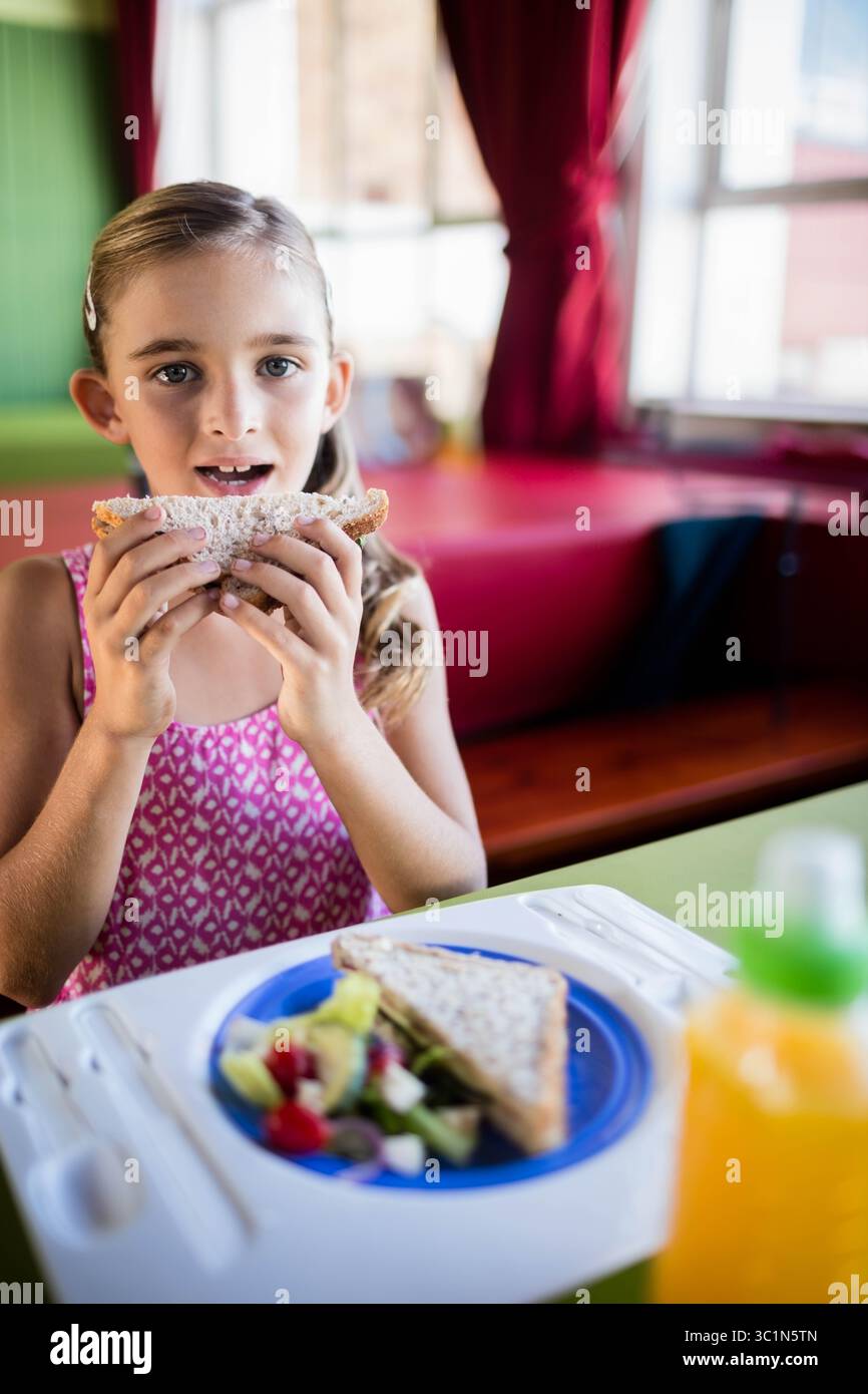 Kind am Stand mit dreieckigem Sandwich in Mundnähe mit Salat auf blauem Teller und Orangensaft Stockfoto
