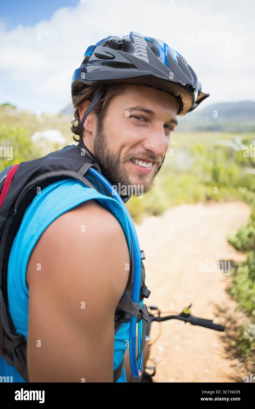 Männlicher Mountainbiker, der auf dem Feldweg steht und mit Helm und Hydratationspack zurückblickt Stockfoto