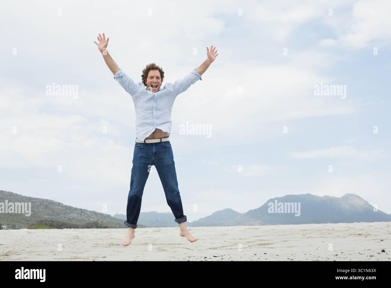 Mittelgroßer Mann, der barfuß auf der Sandküste springt, trägt ein hellblaues Hemd und eine dunkle Jeans Stockfoto