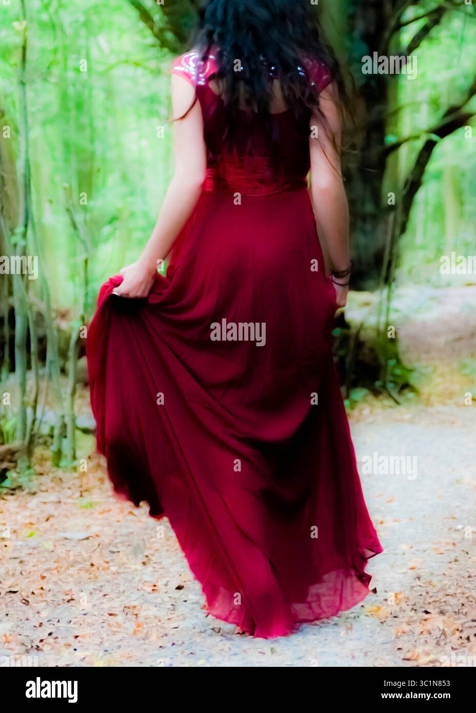 Eine Frau in einem langen roten Kleid geht von der Kamera weg auf einem Weg in einem Wald. Stockfoto