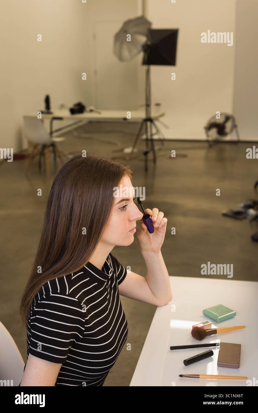 Frau, die Mascara am weißen Tisch im Studio mit Kompakten, Bürsten und Schirmlampe aufträgt Stockfoto
