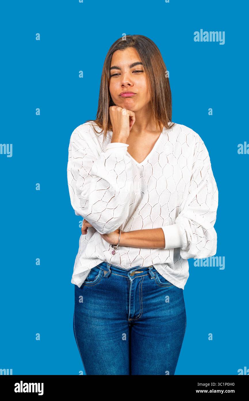 Nachdenkliche junge Frau, die ihr Kinn hält, in Gedanken verloren vor einem leuchtend blauen Hintergrund Stockfoto
