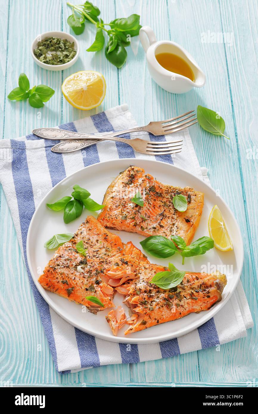 Gegrillte ganze Lachsfilets mit Haut auf einem Teller mit frischem Basilikum, Zitronenscheiben, Gabel und Gewürzen auf Holztisch, vertikale Ansicht von oben, keine Stockfoto