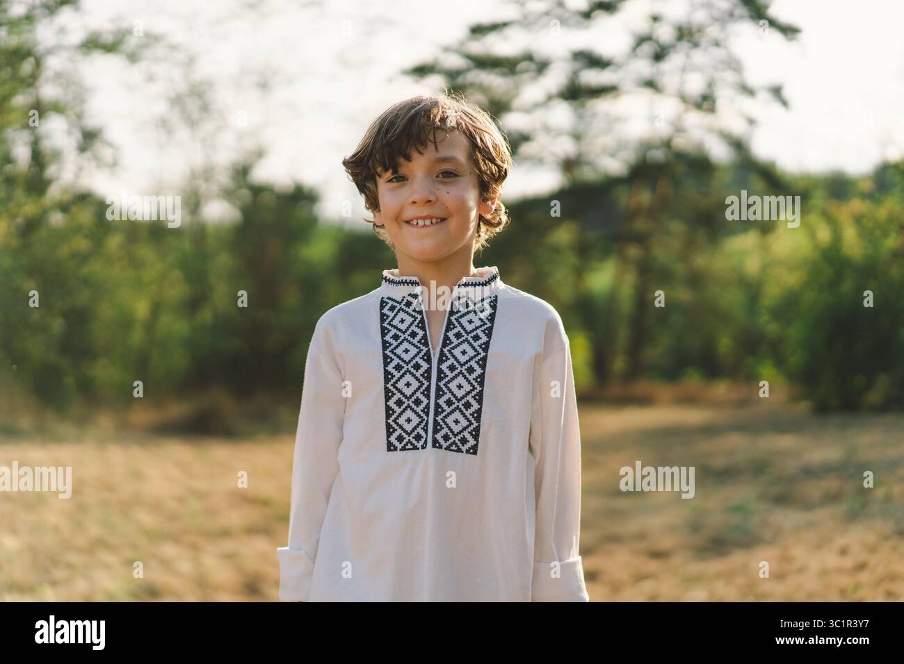 Ein Junge lächelt selbstbewusst, während er auf einem sonnendurchfluteten Feld steht und ein traditionelles besticktes Hemd trägt Stockfoto