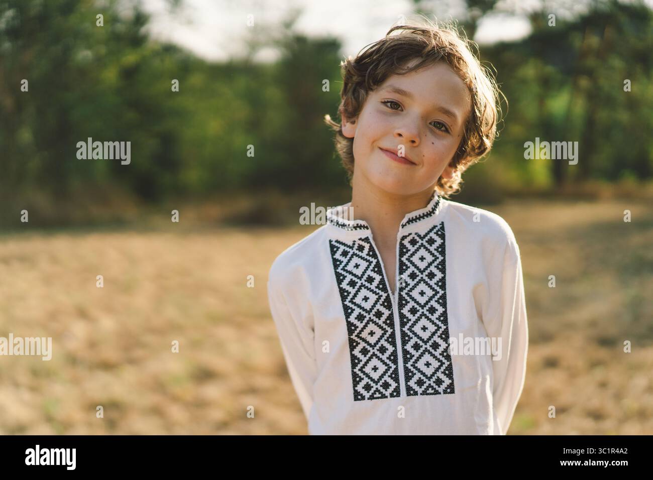 Ein Junge lächelt selbstbewusst, während er auf einem sonnendurchfluteten Feld steht und ein traditionelles besticktes Hemd trägt Stockfoto