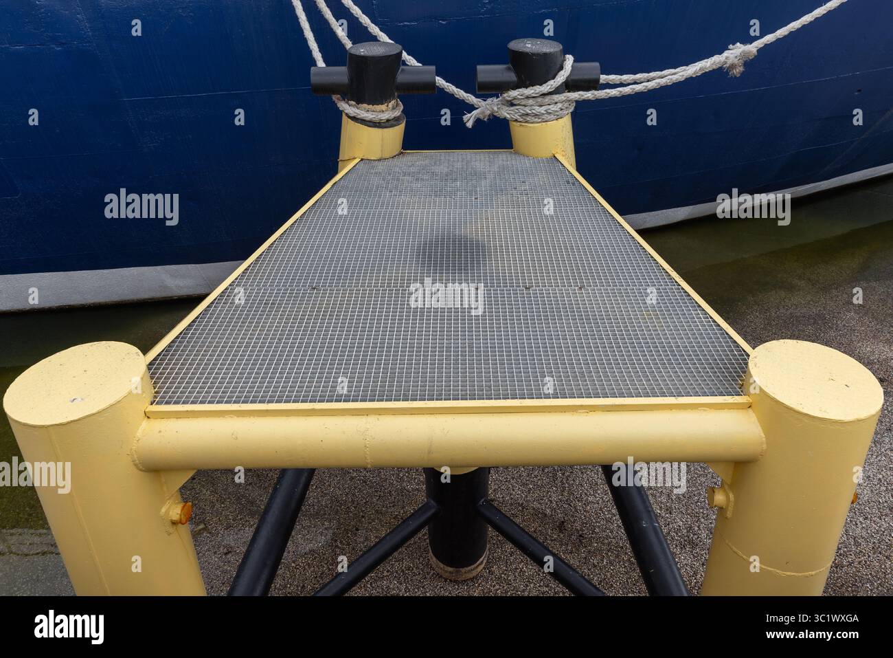 Eine stabile Metallplattform an einem Dock, verbunden mit einem Schiff mit Seilen für Stabilität. Der lebhafte Kontrast von Gelb und Blau zeigt eine maritime Umgebung Stockfoto