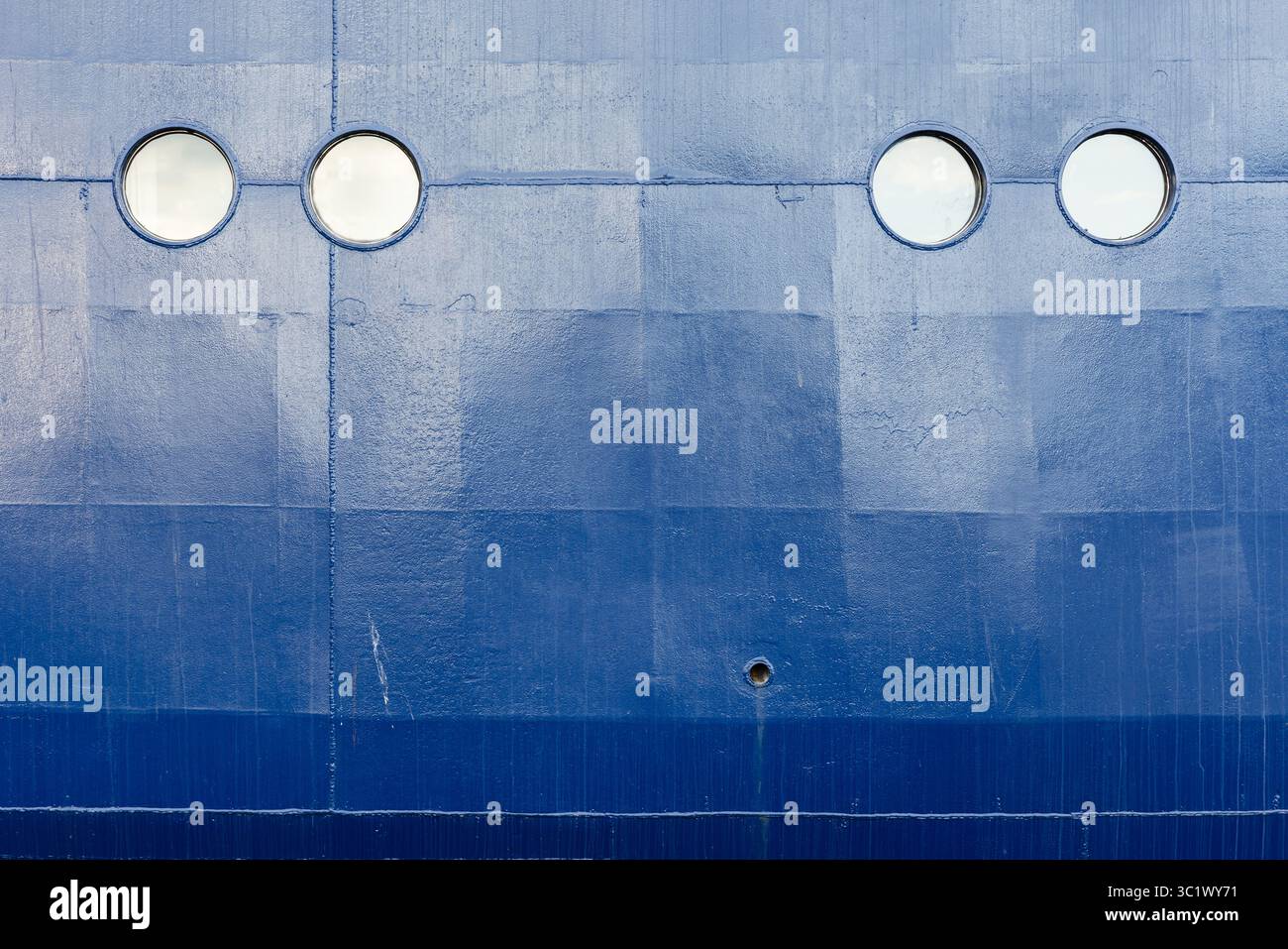 Nahaufnahme eines blauen Schiffrumpfes mit vier kreisförmigen Bullaugen, die in einer geraden Reihe angeordnet sind. Die strukturierte Metalloberfläche und die dezenten Kontraste betonen Stockfoto